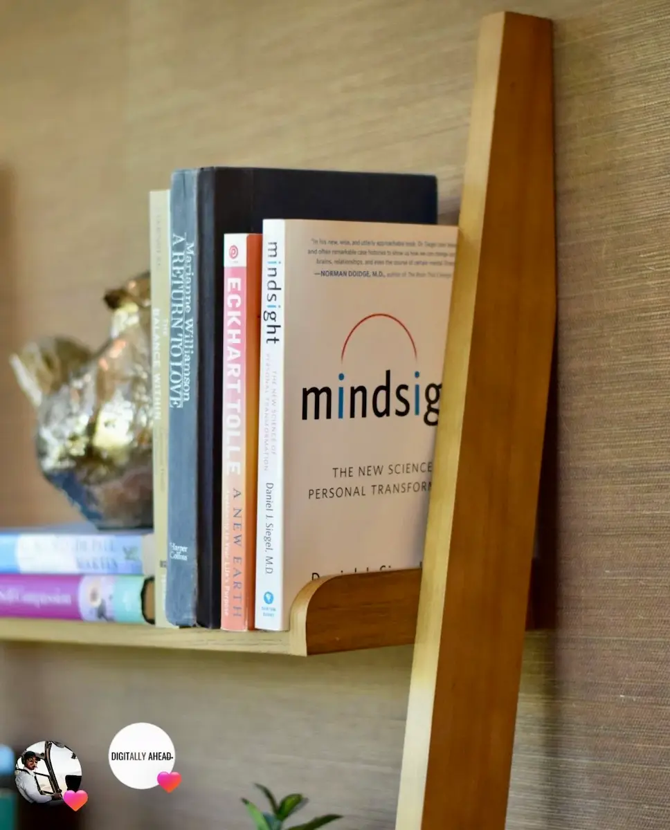 Wooden shelf mounted on a textured beige wall holding several books, including "Mindsight: The New Science of Personal Transformation" by Daniel J. Siegel and "A New Earth" by Eckhart Tolle.