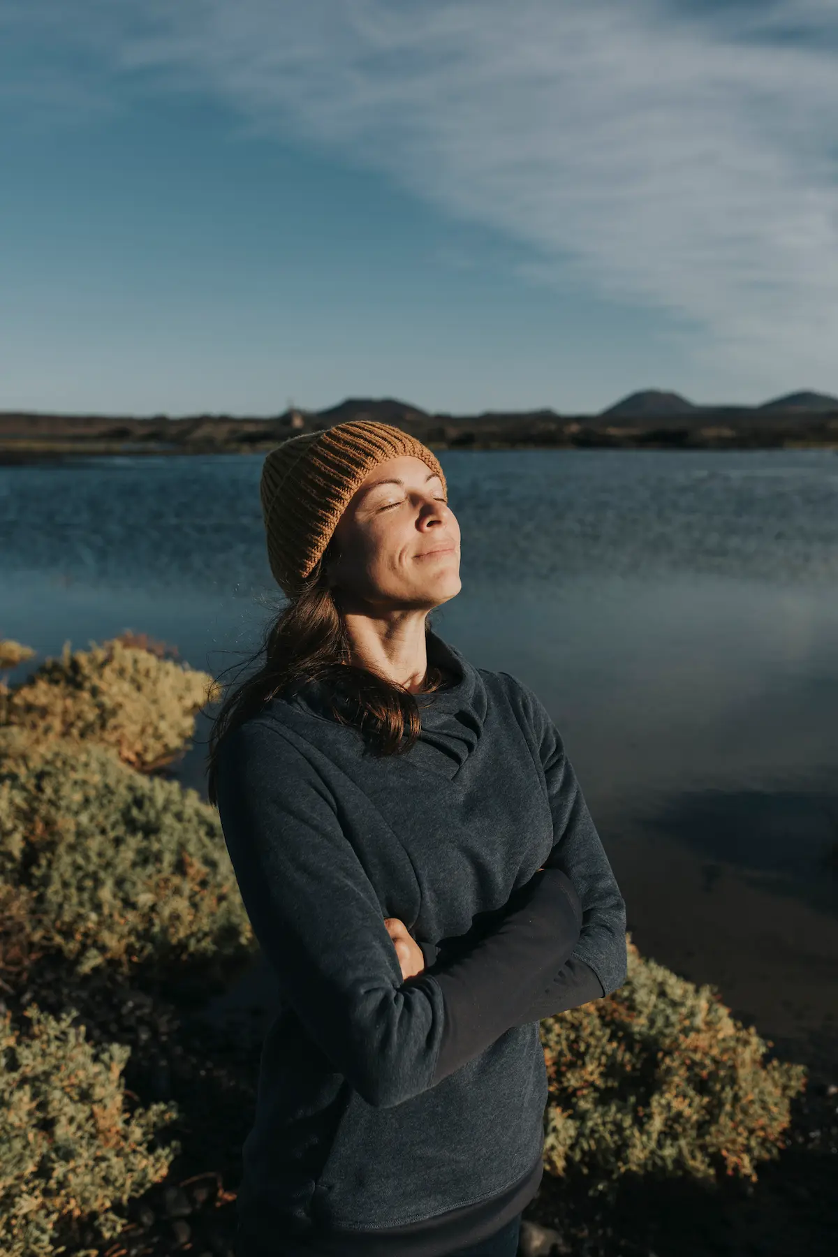 Woman wearing a brown knit hat and dark sweater standing with arms crossed by a lakeside with shrubs, eyes closed and face turned toward the sun under a partly cloudy sky.