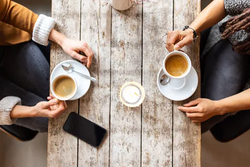 Overhead view of two people sitting across a wooden table, each holding a cup of coffee with saucers and spoons, with a smartphone on the table between them.