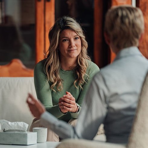 Young woman with long blonde hair in a green sweater sitting on a couch attentively listening to another person in a light-colored shirt during a conversation indoors.