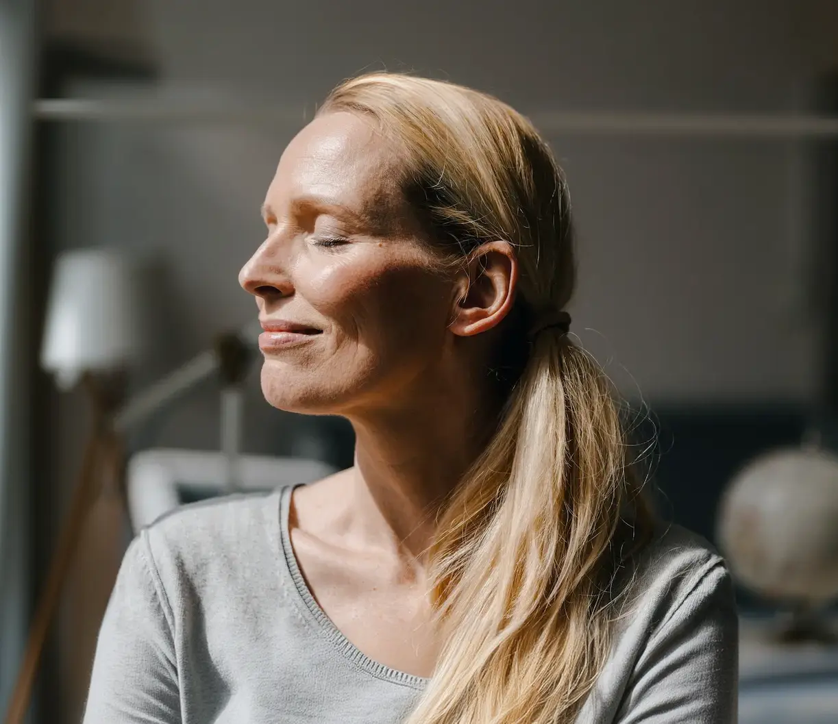 Blonde woman with her hair tied back in a ponytail, eyes closed, and a peaceful expression, sitting indoors with soft lighting.