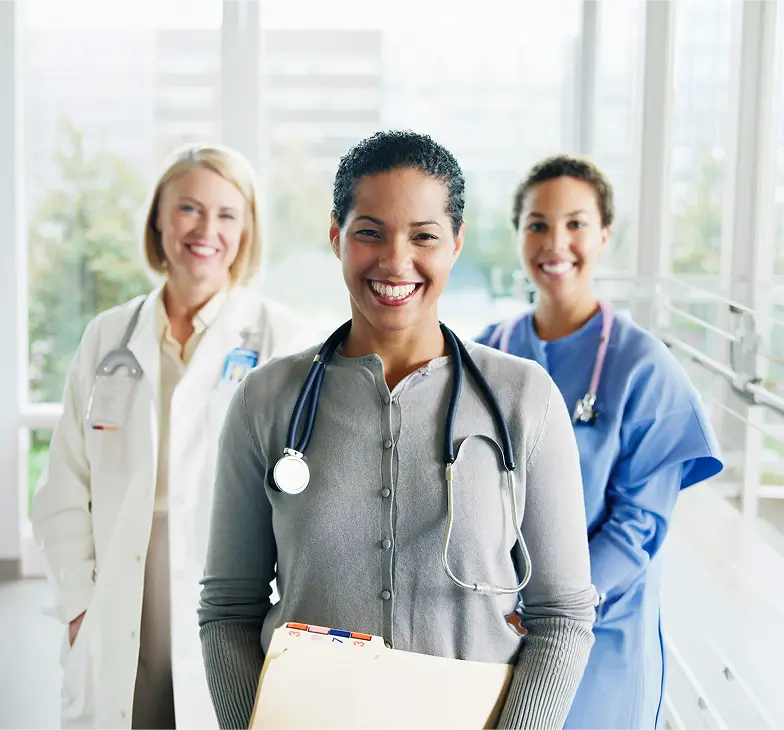 Three smiling female healthcare professionals standing in a bright hospital corridor, one in the front holding a folder and wearing a stethoscope, with two others in medical coats and scrubs behind her.