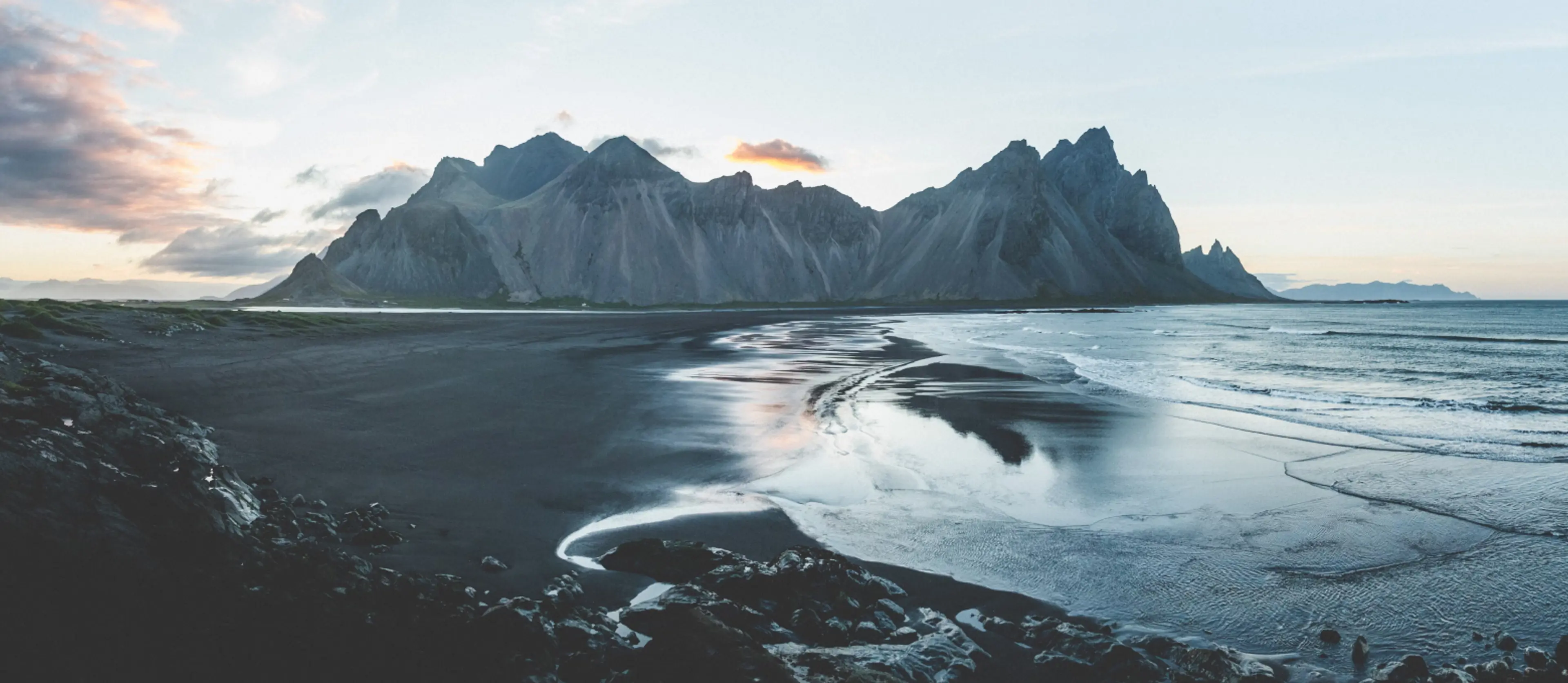 Black sand beach and rocky shore with tall jagged mountains under a cloudy sky at dusk.