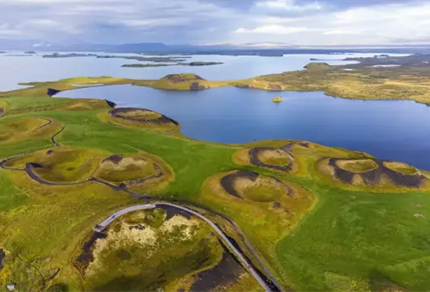 Aerial view of green land with volcanic pseudo craters surrounding a blue lake under a cloudy sky.