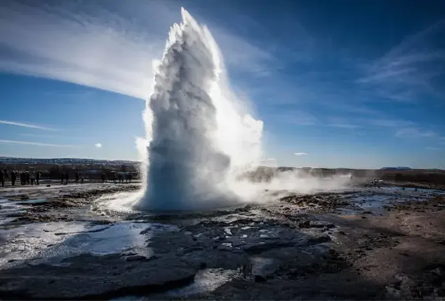 Geyser erupting water and steam high into clear blue sky over rocky ground.