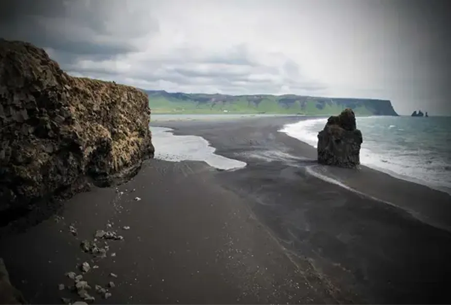 Black sand beach with large rock formations and green cliffs in the distance under cloudy sky.