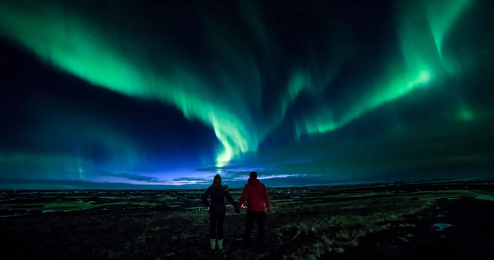 Two people holding hands outdoors at night under green and blue northern lights in the sky.
