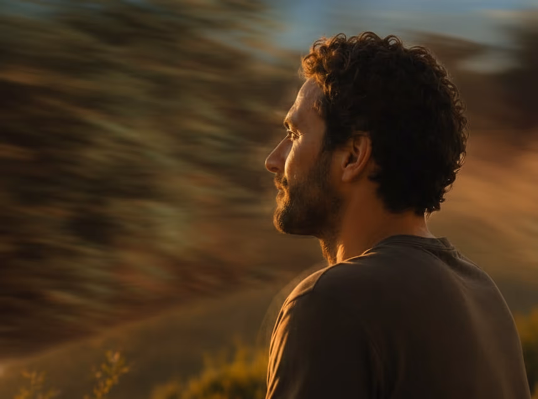 Side profile of a man with curly hair and beard looking thoughtfully into a soft light outdoors.