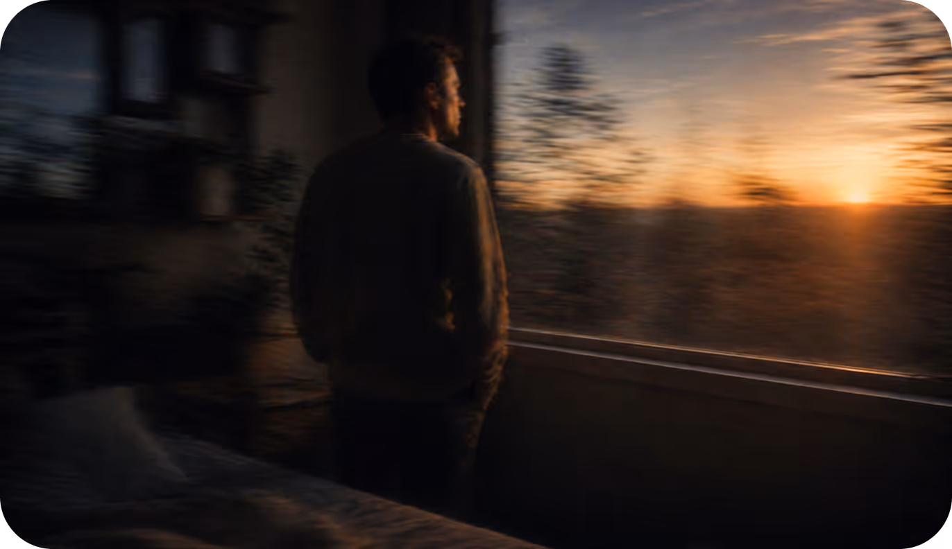 Man standing inside by window looking out at sunset over trees and fields.
