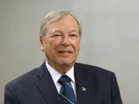 Portrait of an older man in a dark suit and striped tie against a plain gray background.