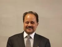 Smiling man in a dark suit, white shirt, and striped tie against a plain background.