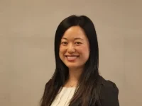 Smiling woman with long dark hair wearing a black blazer and white top standing against a beige wall.