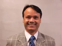 Smiling man in a checkered blazer, blue patterned tie, and white shirt against a plain background.