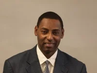 Portrait of a man in a dark pinstripe suit and tie against a plain beige background.