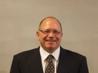 Smiling man with glasses wearing a dark suit, white shirt, and patterned tie against a plain background.