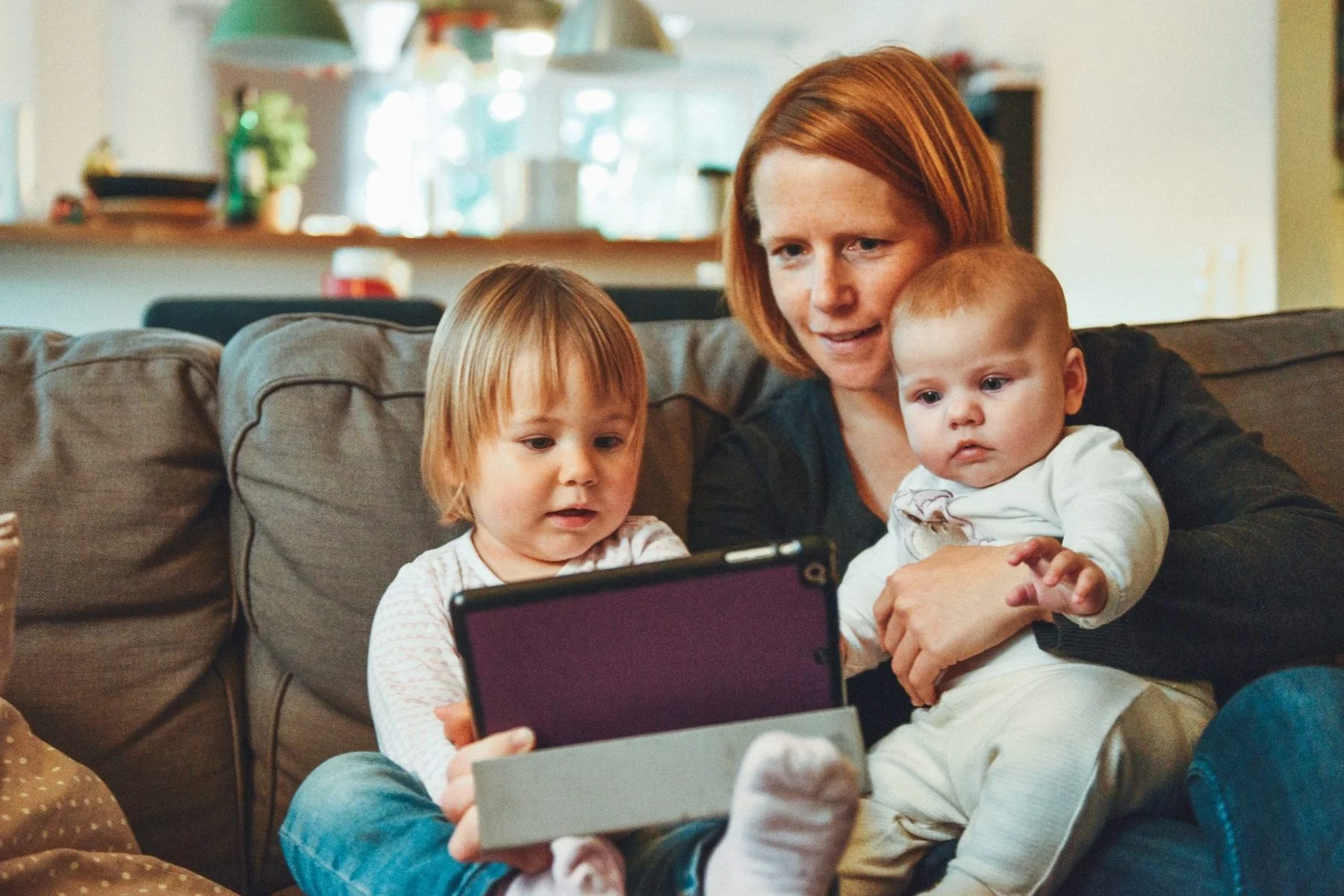 Woman sitting on couch with two young children, all looking at a tablet together.