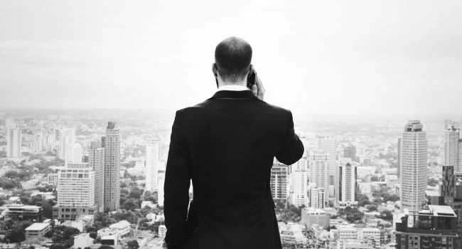 Man in suit talking on phone facing a cityscape with tall buildings and haze in the background.