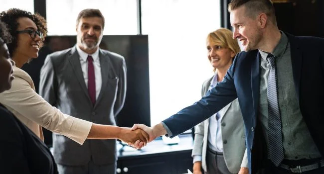 Businesspeople shaking hands in an office with colleagues watching and smiling.