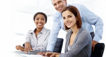 Three diverse business professionals smiling and working together at a desk in an office.