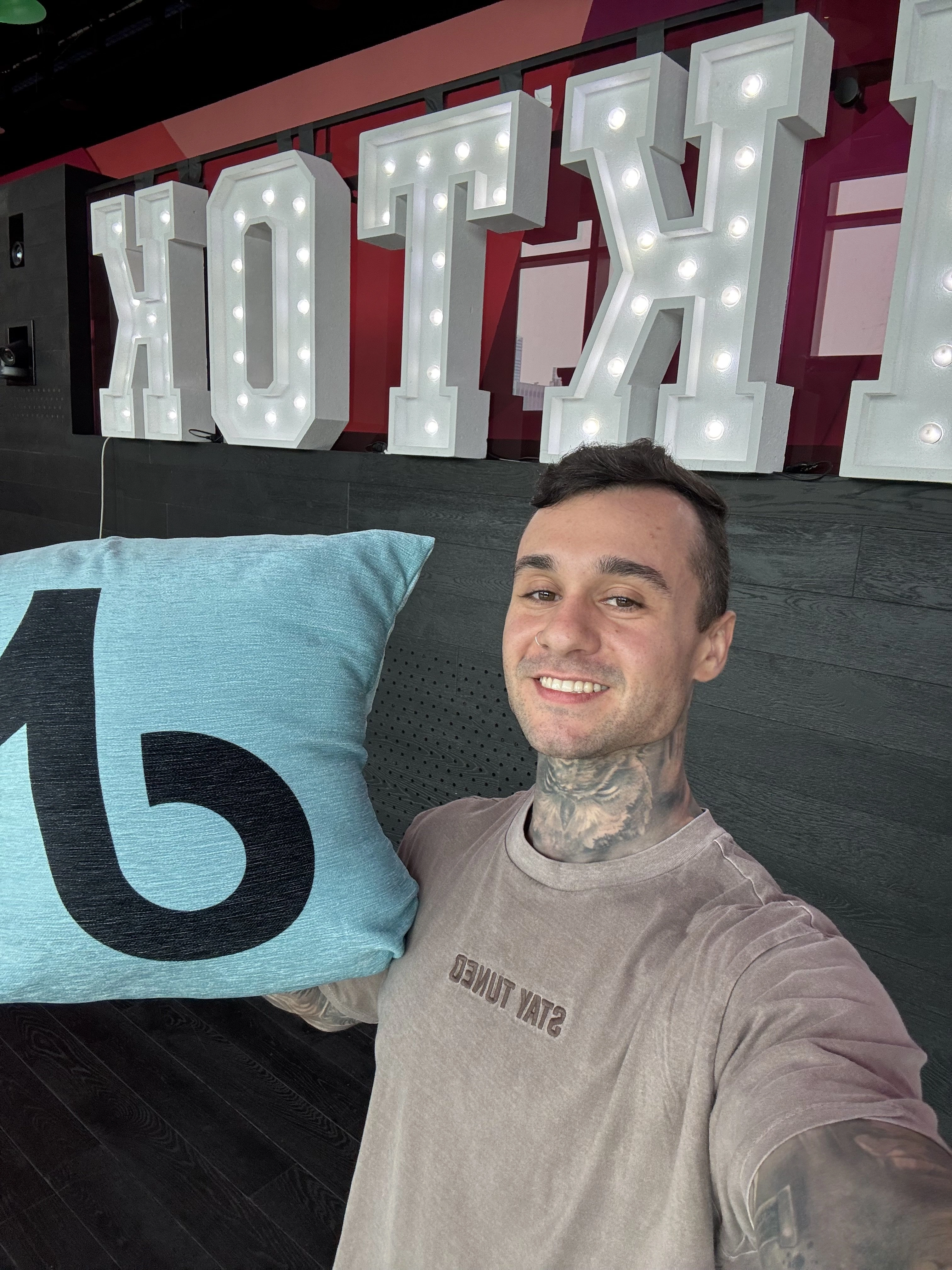 Smiling man with neck tattoos holding a light blue pillow with a black 'b' in front of large lit letters.