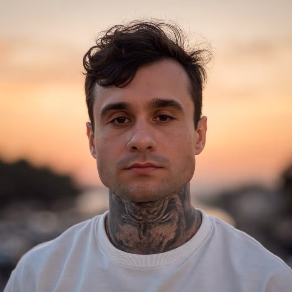 Close-up of a man with a neck tattoo wearing a white shirt against a sunset background.