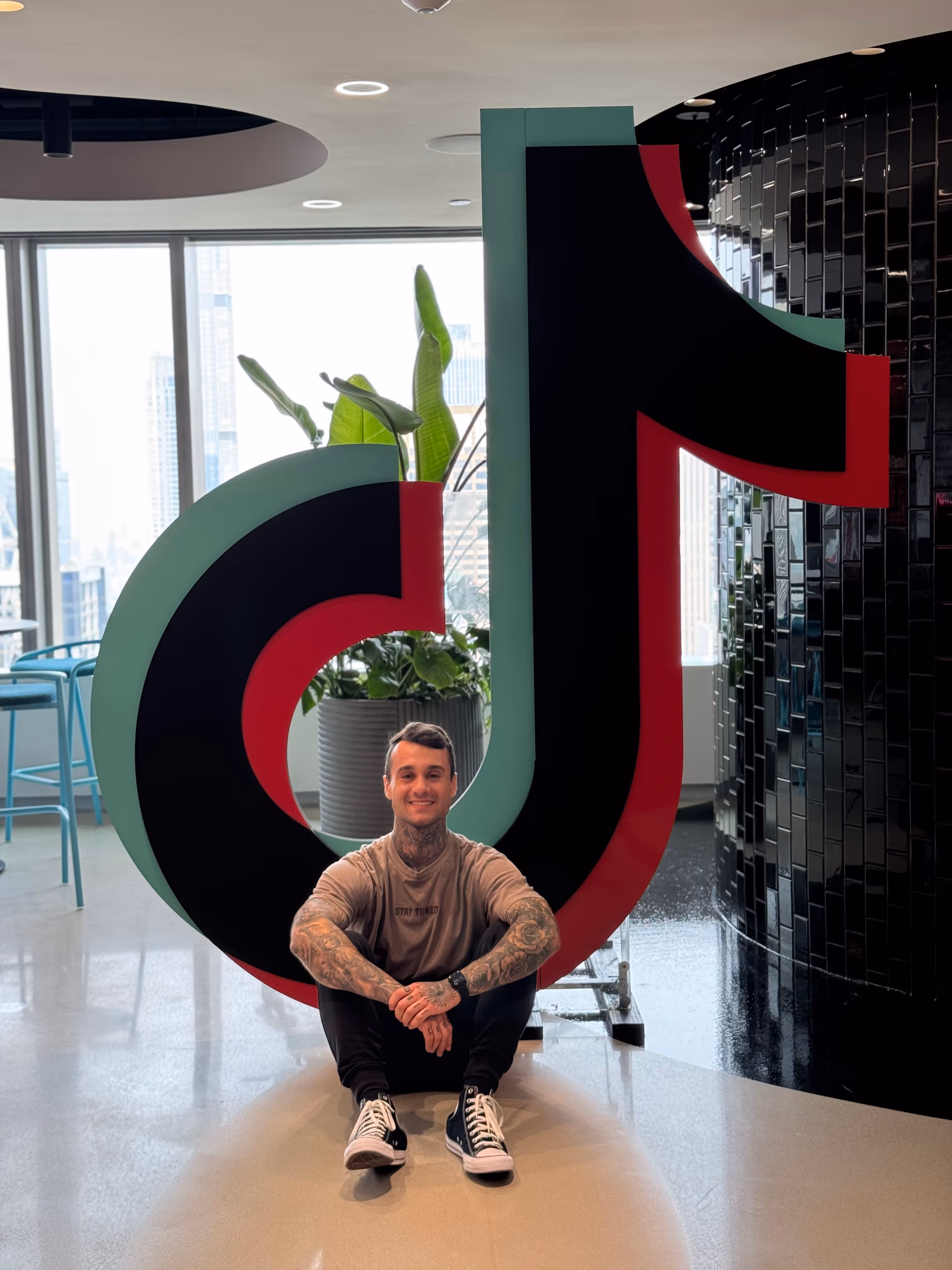 Smiling man with tattoos sitting on floor inside large TikTok logo sculpture in modern indoor space.