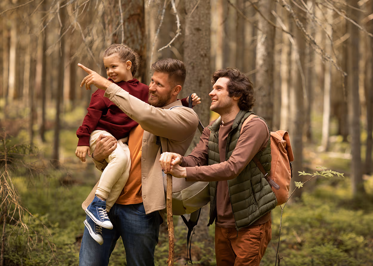 Two men hiking in a forest, one holding a child pointing ahead, the other with a walking stick.