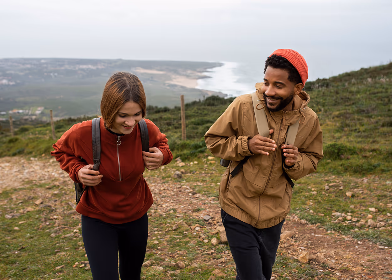 Smiling man and woman hiking uphill with backpacks on a rocky coastal trail.