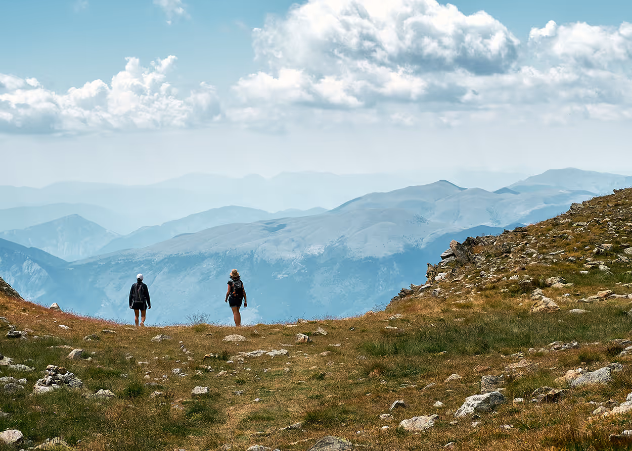 Two hikers walking on a rocky grassy mountain trail with misty mountain ranges and cloudy sky in the distance.