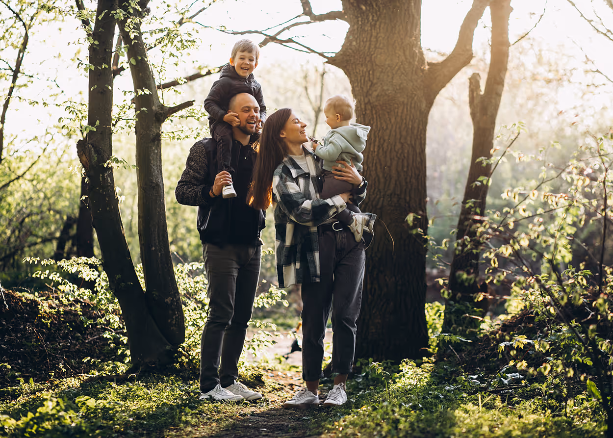 Family of four outdoors: father carries son on shoulders, mother holds toddler in a sunlit forest.