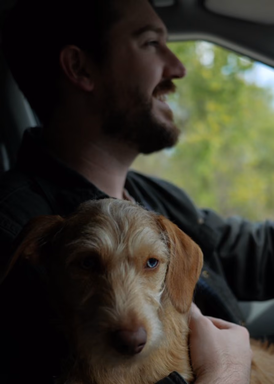 Man with beard smiling while holding a light brown dog with one blue eye inside a car. James Bass, owner of James Bass Counseling in Plano TX