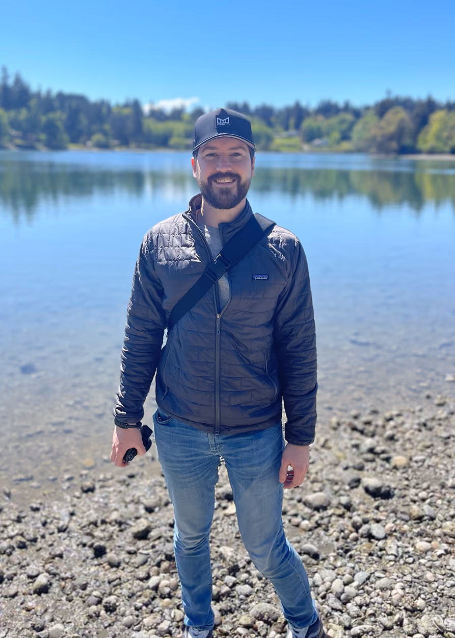 Man wearing a black jacket and cap standing on rocky shore in front of a calm lake with trees in background. James Bass, owner of James Bass Counseling in Plano TX