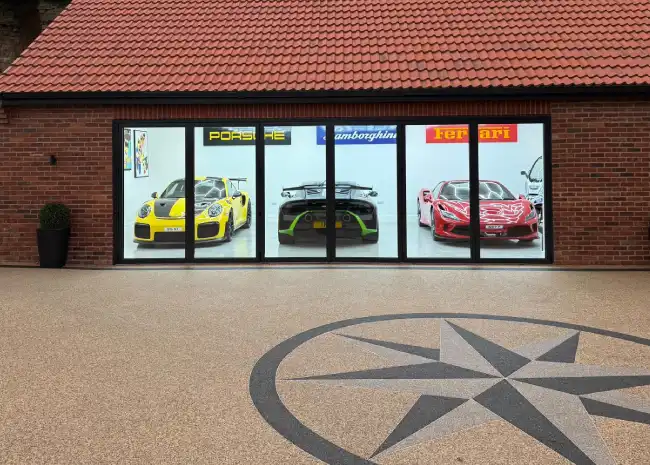 Three luxury sports cars—a yellow Porsche, a black Lamborghini, and a red Ferrari—inside a brick garage in front of a resin bound driveway.