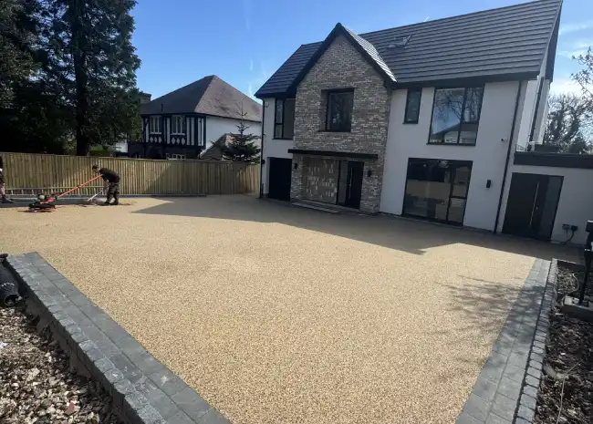 Person leveling freshly laid gravel driveway in front of a modern two-story house with stone and white facade.