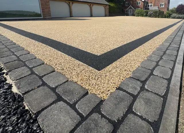 Tan and black resin bound driveway bordered with gray cobblestone pavers and black asphalt.