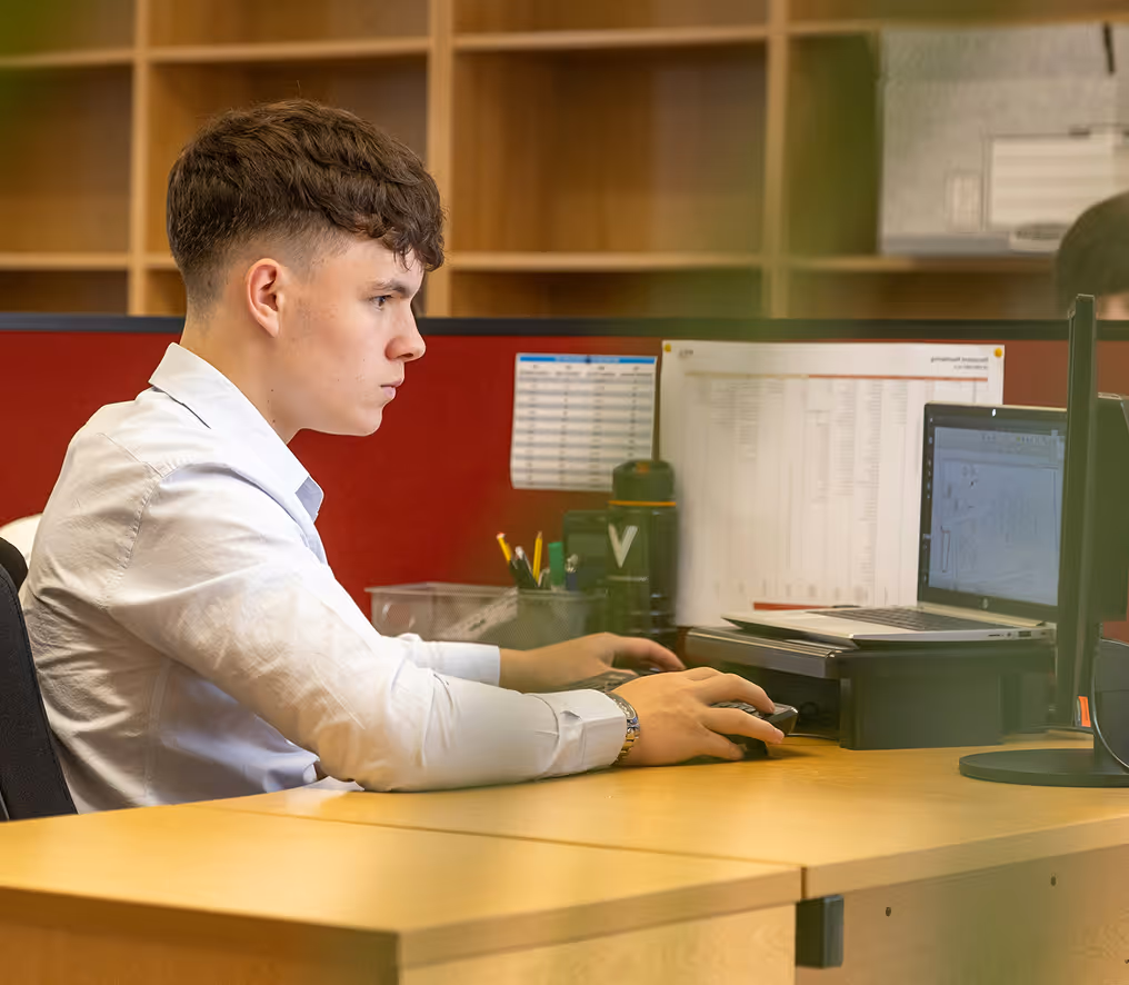 Young man in a white shirt focused on working at a computer desk with dual monitors in an office.