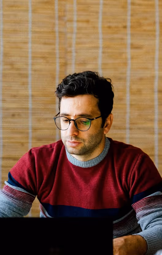 Man with glasses wearing a red and gray striped sweater, looking down at a laptop.