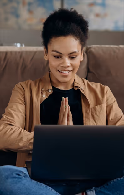 Young woman sitting on a couch with hands pressed together looking at a laptop screen.