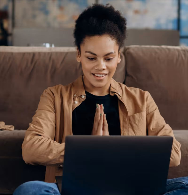 Smiling woman sitting cross-legged on a couch with hands pressed together looking at a laptop screen.