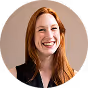 Smiling red-haired woman with long hair wearing a black top against a light brown background.