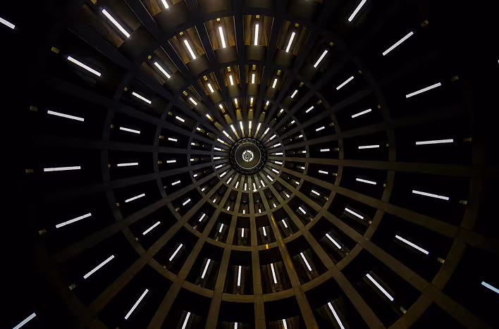 View looking up at a circular, symmetric ceiling structure with radial beams and rectangular light fixtures forming a spiral pattern.