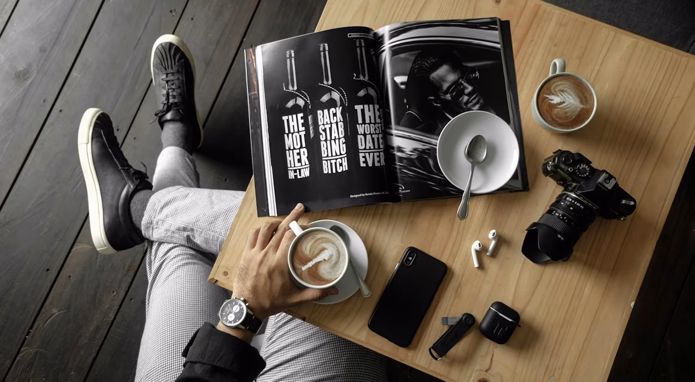 Person sitting with crossed legs at a wooden table holding a cup of latte next to an open book, camera, smartphone, and wireless earbuds.
