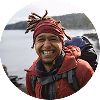 Smiling person wearing a red jacket and headband outdoors with a backpack near a lake and forest.