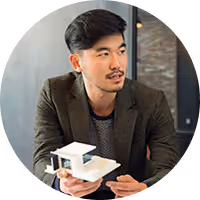 Man with short black hair and a mustache wearing a brown blazer, holding and presenting a small architectural model indoors.