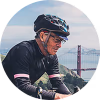 Older man wearing cycling helmet and glasses riding a bike outdoors with the Golden Gate Bridge in the background.