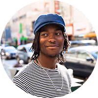 Young man with dreadlocks wearing a navy cap and striped shirt smiling on a city street.