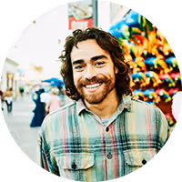 Smiling man with curly hair and a beard wearing a plaid shirt at a colorful outdoor fair.
