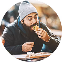 Man wearing a gray beanie and dark jacket eating a sandwich indoors.