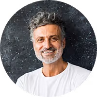 Smiling middle-aged man with curly gray hair and beard wearing a white shirt against a dark speckled background.
