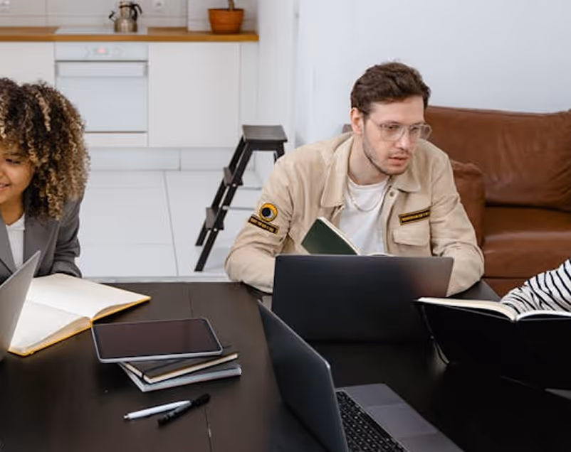 Three young people sitting around a dark table with laptops and books, engaged in studying or discussion in a modern kitchen setting.